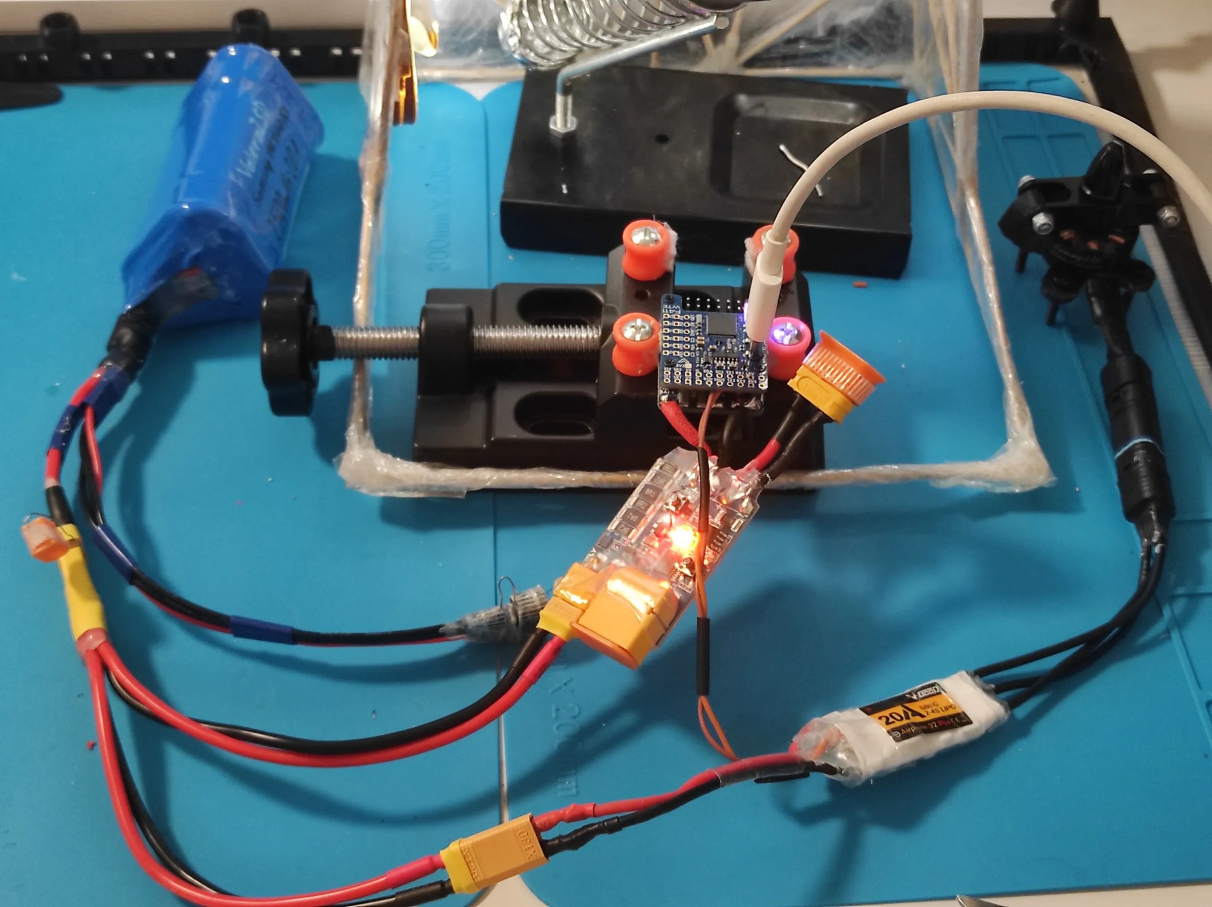 a blue flight controller with a lit status LED in a small vise, wired to a white ESC, a brushless motor, a blue battery pack and an inline XT60 module, on a blue soldering mat