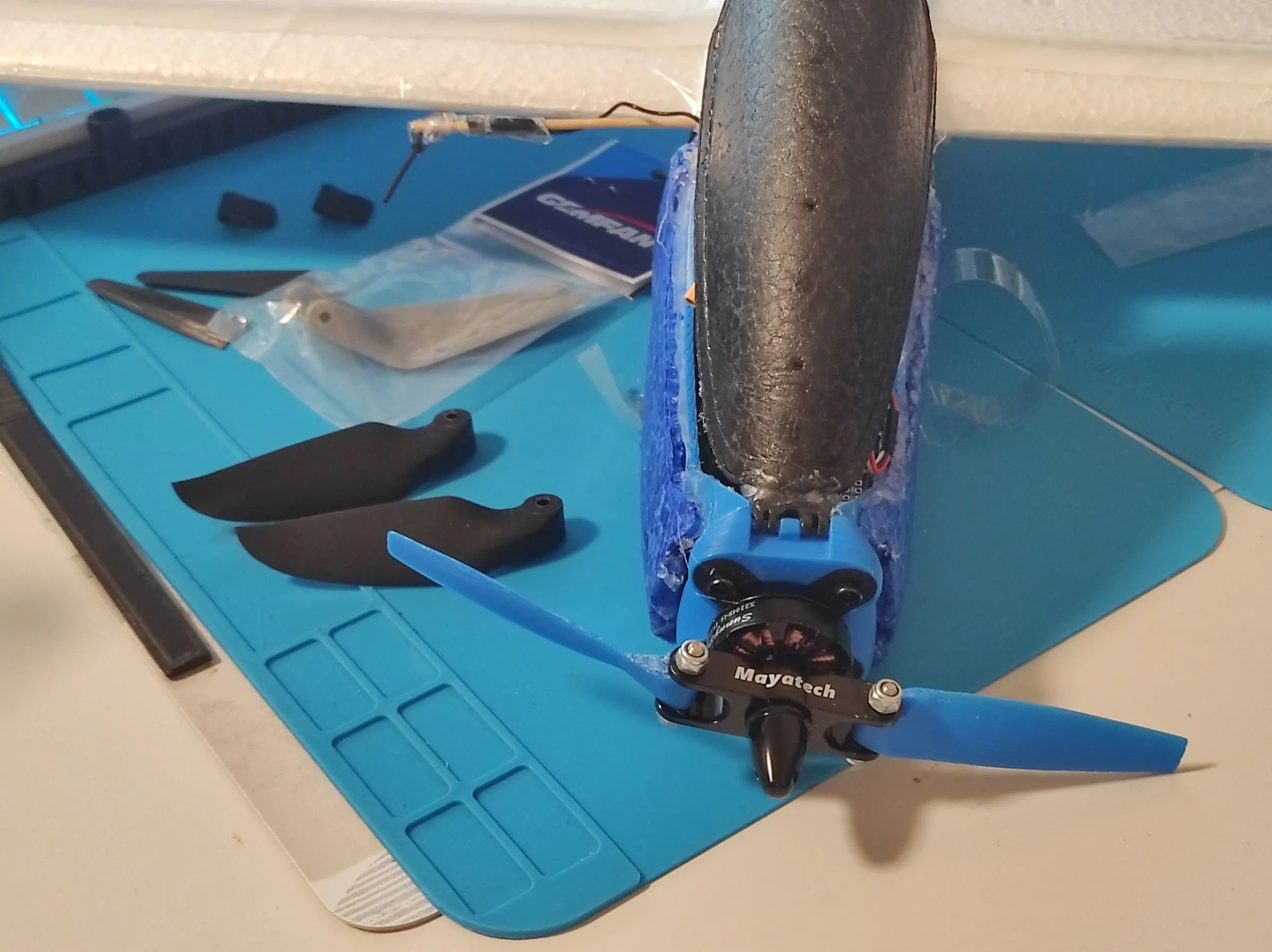 the nose of the foam aircraft with blue printed folding blades on a Mayatech hub, two black printed blades and a Gemfan bag with grey moulded blades on a blue mat beside it