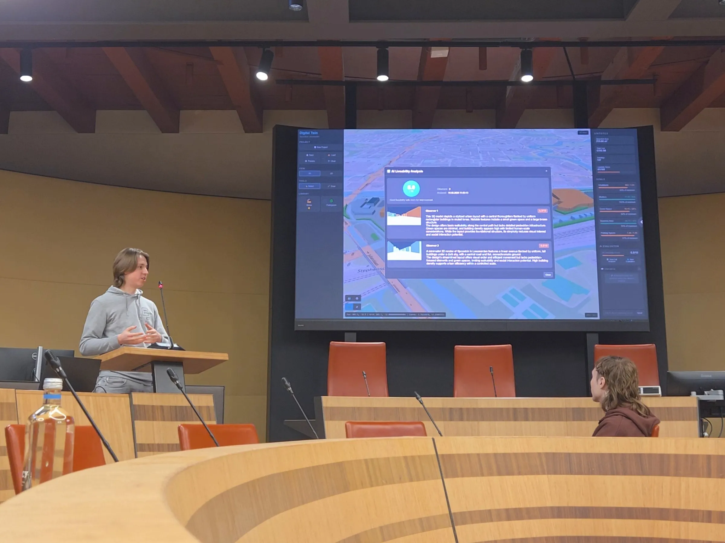 Valerii at a lectern in the council chamber, beside a large screen showing the application's AI liveability analysis over the 3D city